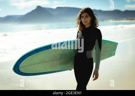 Porträt einer Frau mit gemischter Rasse, die an einem sonnigen Tag am Strand Surfbrett hält Stockfoto