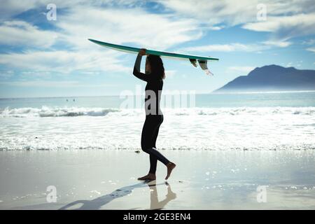 Frau mit gemischtem Rennen, die an einem sonnigen Tag am Strand Surfbrett hält Stockfoto