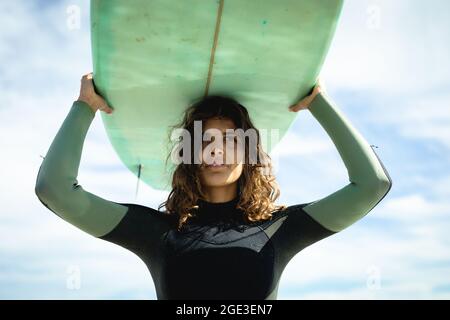 Frau mit gemischtem Rennen, die an einem sonnigen Tag am Strand Surfbrett hält Stockfoto