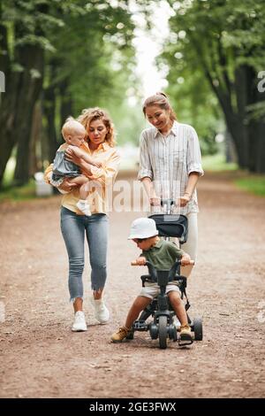 Vertikale Ganzkörperportrait von zwei jungen Müttern, die mit Babys im Park spazieren und sich unterhalten Stockfoto