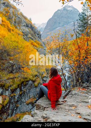 Felsige Berge und herbstliche Herbstbäume und Touristenfrau sitzen auf Felsen. Berglandschaft mit Fluss Stockfoto