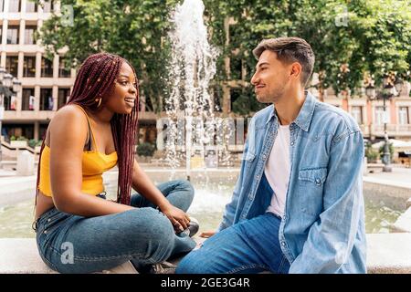 Multikulturelle junge Freunde, die neben einem Brunnen sitzen und sich unterhalten und angusten. Stockfoto