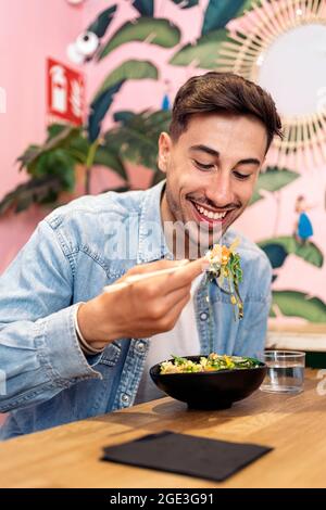 Junger Mann, der in einem Restaurant sitzt und einen gesunden Poke genießt und Essstäbchen verwendet. Stockfoto