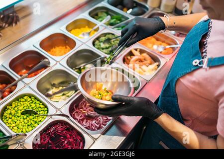 Nicht anerkannter Restaurantmitarbeiter in Schutzhandschuhen, der Zutaten in eine Poke-Schüssel gegeben hat. Stockfoto