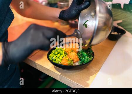 Nicht anerkannter Restaurantmitarbeiter in Schutzhandschuhen, der Zutaten in eine Poke-Schüssel gegeben hat. Stockfoto