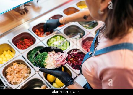 Nicht anerkannter Restaurantmitarbeiter mit Schutzhandschuhen, der Zutaten in eine Schüssel mit Reis und Gemüse steckt. Stockfoto
