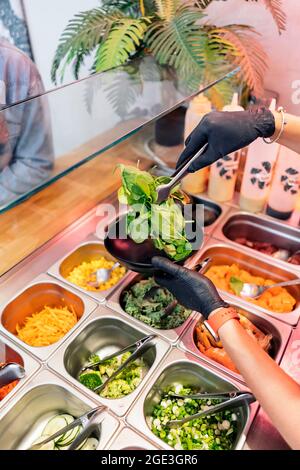 Nicht anerkannter Restaurantmitarbeiter in Schutzhandschuhen, der Gemüse in die Schüssel einfügte. Stockfoto