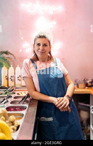 Blonde Frau mit Schürze lächelnd und Blick auf die Kamera während der Arbeit in einem Restaurant. Stockfoto