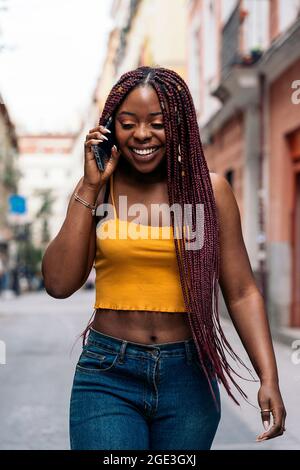Fröhliche junge afrikanische Frau mit Zöpfen, die auf der Straße gehen und am Telefon sprechen. Stockfoto