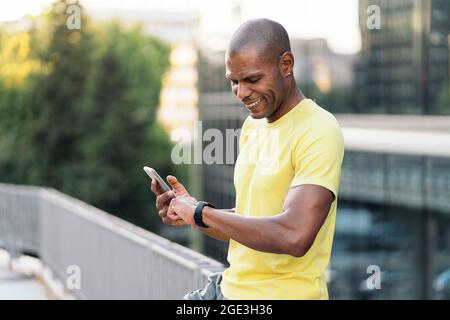 Fröhlicher afroamerikanischer Sportler mit gelbem T-Shirt, das Handy und Smartwatch synchronisiert, um in der Stadt Sport zu treiben. Stockfoto