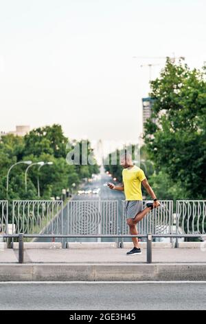 Afroamerikanischer Mann, der sein linkes Bein streckte und am Tag das Telefon auf dem Steg hielt. Stockfoto