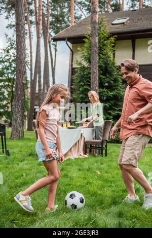 Lächelndes Kind spielt Fußball mit Vater in der Nähe der Mutter und am Tisch im Freien Stockfoto