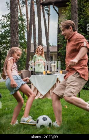 Lächelnde Frau mit Teller, der neben dem Tisch stand, und verschwommener Familie, die Fußball spielt Stockfoto