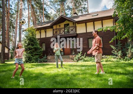 Positive Familie spielt Volleyball auf Rasen in der Nähe des Ferienhauses Stockfoto