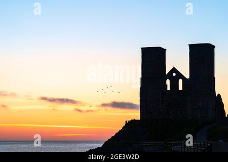 Der Morgenhimmel mit einer kleinen Vogelschar, die über dem Meer fliegt, und die Silhouette der angelsächsischen Zwillingstürme der Reculver-Kirche aus dem 11. Jahrhundert. Stockfoto