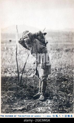 Frühe japanische Postkarte eines Eingeborenen, Ainu, aus Hokkaido, Nordjapan. Reifer Mann mit typischem Ainu-Bart, der mit Bärenjungen auf dem Rücken steht Stockfoto