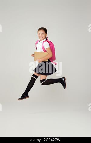 Nettes Schulmädchen springen mit rosa Rucksack isoliert auf grauem Hintergrund. Glückliches Kind ist bereit zu lernen. Zurück zur Schule Stockfoto