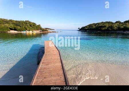 Wunderschönes Ionisches Meer mit klarem türkisfarbenem Wasser, hölzernen Pier und Blick auf die Küste mit feinem Sand vom Ksamil Strand in Albanien Stockfoto