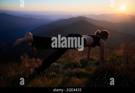 Junge Frau in schwarzer Sportkleidung, die bei Sonnenuntergang spezielle Yoga-Übungen zur Natur in den Bergen macht. Konzept der Yoga-Zeit. Stockfoto