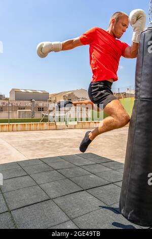 Der kaukasische Youong-Mann trifft mit dem Knie einen Boxsack, während er Kikcboxing trainiert. Machen Sie einen spektakulären Sprung nach oben zu treffen. Unterhaltung Stockfoto