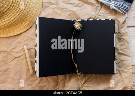 Flache Lay-Komposition mit einem offenen leeren schwarzen Notizbuch, Bleistift, schwarz-weißer Postkarte, getrocknetem Lavendel und Strohhut auf Bastelpapier-Hintergrund. Draufsicht. Stockfoto