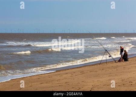 Menschen Seefischen am Sandstrand von Chapel St Leonards in Lincolnshire, England, mit Offshore-Windpark-Turbinen am Horizont sichtbar. Stockfoto