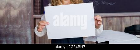 Die Schule ist ein glücklicher Ort. Glückliches kleines Kind, das leeres pinkfarbenes Papier auf gelbem Hintergrund hält. Kleines Mädchen lächelt glücklich mit leerem Plakat. Es wird Jo Stockfoto