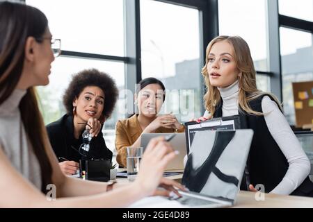 Verschwommene Geschäftsfrau, die auf einen Laptop zeigt, während sie mit Kollegen aus verschiedenen ethnischen Gruppen zusammenarbeitet Stockfoto