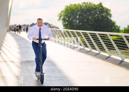 Ein lustiger Geschäftsmann genießt seine Fahrt mit einem Elektroroller, als er bei Sonnenuntergang eine lange Brücke überquert und dabei den Fußgängerweg in einem städtischen Nahverkehr benutzt Stockfoto