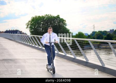 Ein lustiger Geschäftsmann genießt seine Fahrt mit einem Elektroroller, als er bei Sonnenuntergang eine lange Brücke überquert und dabei den Fußgängerweg in einem städtischen Nahverkehr benutzt Stockfoto