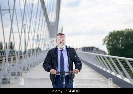 Lächelnder, freundlicher Geschäftsmann, der auf einem Elektroroller fährt, während er bei Sonnenuntergang über eine lange Brücke über einen Fluss nach Hause pendelt Stockfoto