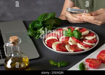 Die junge Frau machte einen Caprese-Salat nach einem Rezept aus dem Internet und fotografiert das Ergebnis der Arbeit mit dem Telefon. Stockfoto