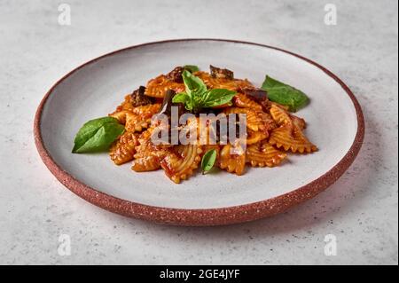 Pasta Farfalle mit Auberginen, Tomatensauce, Käse und Basilikum auf dem Teller auf einem grauen strukturierten Hintergrund, Nahaufnahme Stockfoto