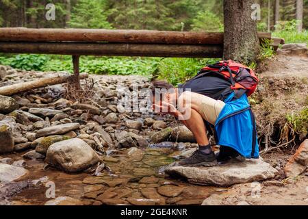 Wanderer mit Rucksack wäscht Gesicht in Bergfluss im Karpaten Wald durch Brücke. Tourist mit Ruhe durch Strom umgeben von Sommernatur Stockfoto