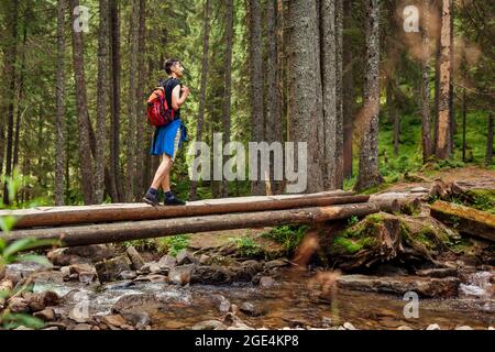 Wanderwanderer mit Rucksack überqueren Bergfluss im Karpaten Wald zu Fuß auf Holzbrücke. Mann Tourist genießen Landschaft. Ausflug zum Sommer Ukr Stockfoto