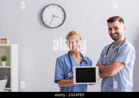 Fröhliche Krankenschwester zeigt digitales Tablet mit leerem Bildschirm in der Nähe eines positiven Arztes, der mit gekreuzten Armen steht Stockfoto