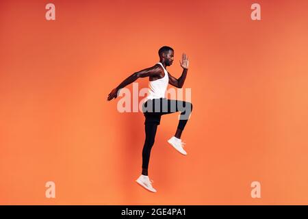 Fit Mann springen im Studio. Männlicher Laufer, der sich vor dem Training vor orangefarbenem Hintergrund aufwärmt Stockfoto