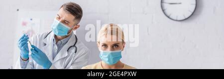Frau in medizinischer Maske, die Kamera ansieht, während Arzt Spritze mit Impfstoff, Banner füllt Stockfoto