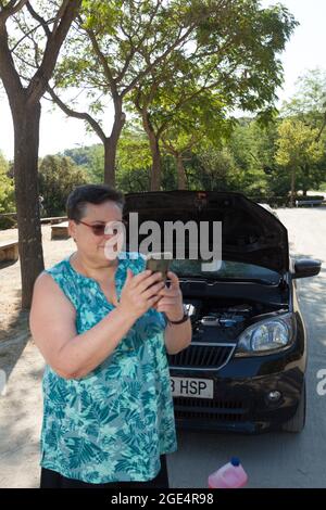 Frau, die mit dem Mobiltelefon die Pannenhilfe vor einem offenen Auto mit Motor in Sicht anruft Stockfoto