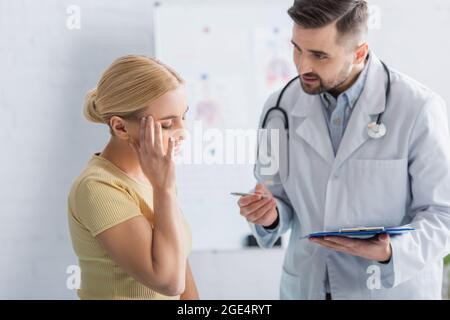 Arzt mit Klemmbrett im Gespräch mit Frau, die den Kopf mit geschlossenen Augen berührt Stockfoto