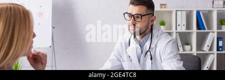 Arzt in weißem Mantel und Brille in der Nähe von blonden Patienten in der Klinik, Banner Stockfoto