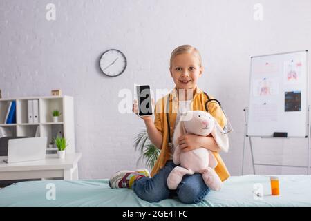 Glückliches Mädchen, das auf einer medizinischen Couch mit Spielzeug-Hase sitzt und Smartphone mit leerem Bildschirm zeigt Stockfoto