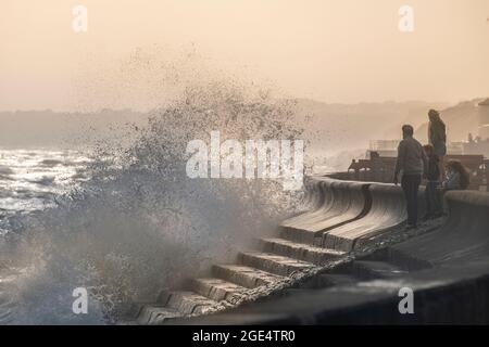 Die Menschen trotzen den rauhen Wellen, die im Sommer auf die Verteidigung des Meeres in Milford-on-Sea in Hampshire stürzen, mit weit entfernten Klippen im Sonnenlicht. Stockfoto