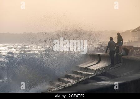 Die Menschen trotzen den rauhen Wellen, die im Sommer auf die Verteidigung des Meeres in Milford-on-Sea in Hampshire stürzen, mit weit entfernten Klippen im Sonnenlicht. Stockfoto