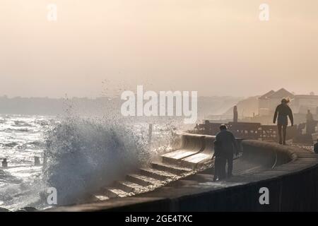 Die Menschen trotzen den rauhen Wellen, die im Sommer auf die Verteidigung des Meeres in Milford-on-Sea in Hampshire stürzen, mit weit entfernten Klippen im Sonnenlicht. Stockfoto