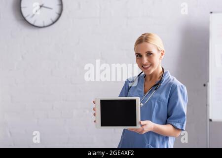 Lächelnder Arzt in blauer Uniform, der ein digitales Tablet mit leerem Bildschirm zeigt Stockfoto