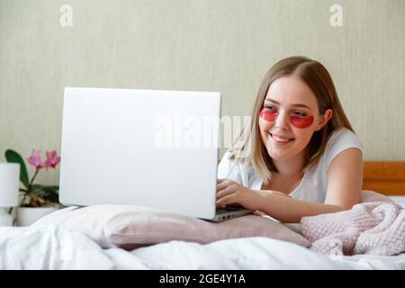 Lächelnde kaukasische Frau im Schlafanzug liegt im Bett und arbeitet mit Laptop. Junge Frau verwendet Hydrogel-Pflaster während der Arbeit. Morgendliche Hautpflege-Rituale Stockfoto