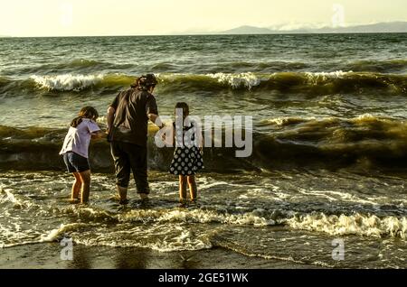 Sevan-See, Armenien, 24. Juni 2021: Eine junge Frau hält zwei kleine Mädchen an den Händen und steht knietief im stürmischen, schaumigen Wasser, beleuchtet von der r Stockfoto