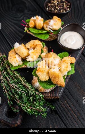 Toasts mit knusprig gebratenen Garnelen und grünem Salat auf einem Holzbrett. Schwarzer Holzhintergrund. Draufsicht Stockfoto