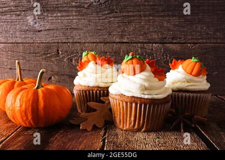 Herbstkürbis würzen Cupcakes mit cremigem Zuckerguss und Herbstüberzügen. Szene vor einem rustikalen Holzhintergrund. Stockfoto